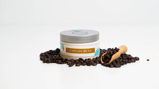 A 4 oz. jar of Cocoa Bean body butter is placed against a white background, encircled by cocoa beans and accompanied by a wooden scooper, highlighting the product's rich, indulgent nature and its key ingredient.
