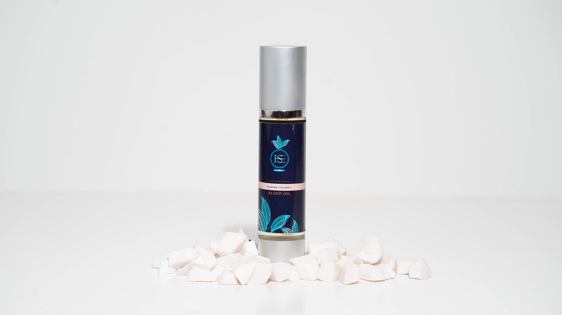 A 30 ml bottle of Almond Coconut Luxe Grooming Oil is centered against a white background, framed by pieces of coconut, highlighting the product's key ingredients and emphasizing its natural, nourishing properties.