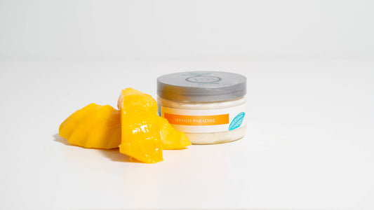 A 4 oz. jar of Mango Paradise body butter is displayed against a white background, accompanied by fresh mango slices placed beside it, highlighting the body butter's tropical mango inspiration and natural ingredients.