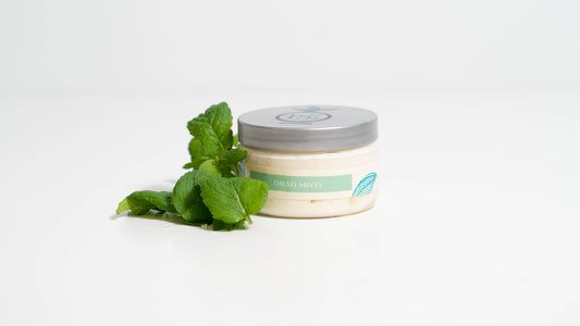 A 4 oz. jar of body butter next to a handful of mint leaves is presented against a white background, indicating a simple, elegant design suitable for highlighting the product's purity and quality.
