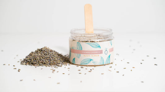 A 16 oz. jar of Calming LaVanilla body scrub is positioned against a white background, featuring a wooden applicator inside and encircled by lavender buds, emphasizing the scrub's soothing lavender and vanilla essence.