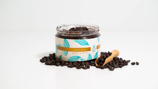 A 16 oz. jar of Cocoa Bean body scrub is presented against a white background, encircled by cocoa beans with a wooden scooper alongside, illustrating the scrub's rich cocoa-infused formula and natural elements.