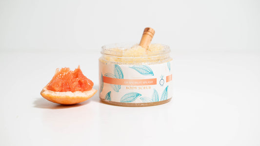 A 16 oz. jar of Grapefruit Splash body scrub is displayed against a white background, featuring a wooden scooper inside the jar and a slice of grapefruit placed beside it, showcasing the scrub's fresh grapefruit ingredient and invigorating scent.