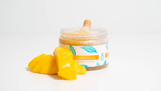A 16 oz. jar of Mango Paradise body scrub is displayed against a white background, accompanied by a wooden scooper inside the jar and fresh mango slices placed beside it, highlighting the scrub's natural mango essence and ingredients.