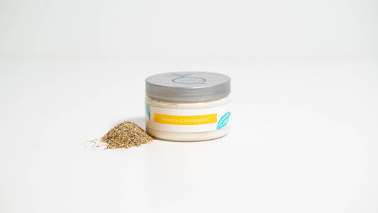 A 4 oz. jar of Vanilla Chamomile body butter is shown against a white background, with dried chamomile flowers placed beside it, conveying the product's soothing essence and natural ingredients.