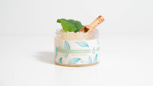 A 16 oz. jar of Oh So Minty body scrub is shown against a white background, decorated with mint leaves and containing a wooden scooper inside, showcasing the scrub's refreshing ingredients and natural appeal.